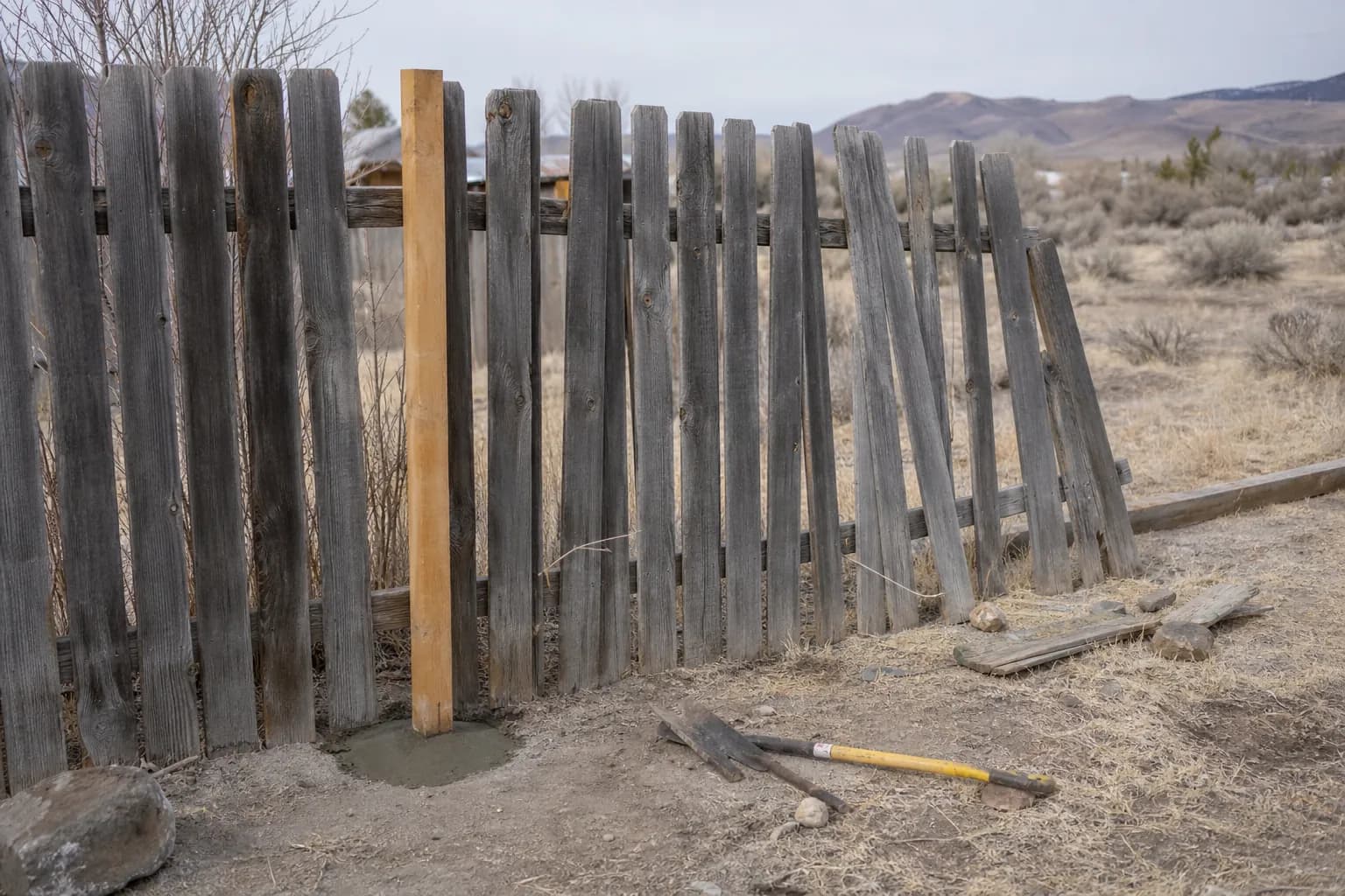 Weathered gray wood fence section leaning after wind with one new cedar post set in fresh concrete